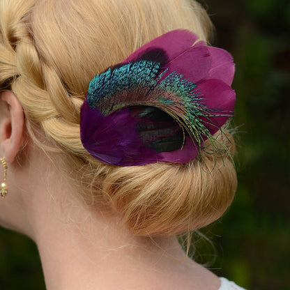 A close-up of a blonde woman's braided bun with a large decorative feather hair accessory in purple and iridescent teal-green, worn in a real outdoor setting with blurred greenery in the background.