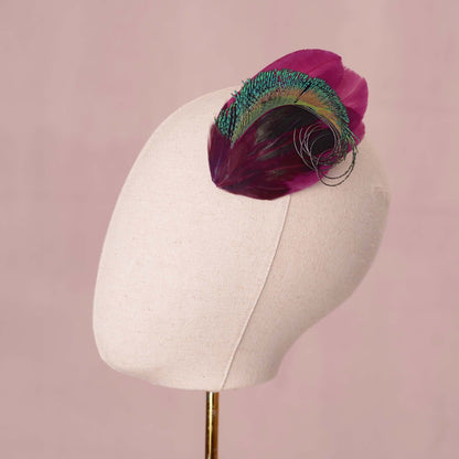A decorative feather hair accessory in rich purple and iridescent green-blue tones, displayed on a cream fabric mannequin head against a soft pink background.