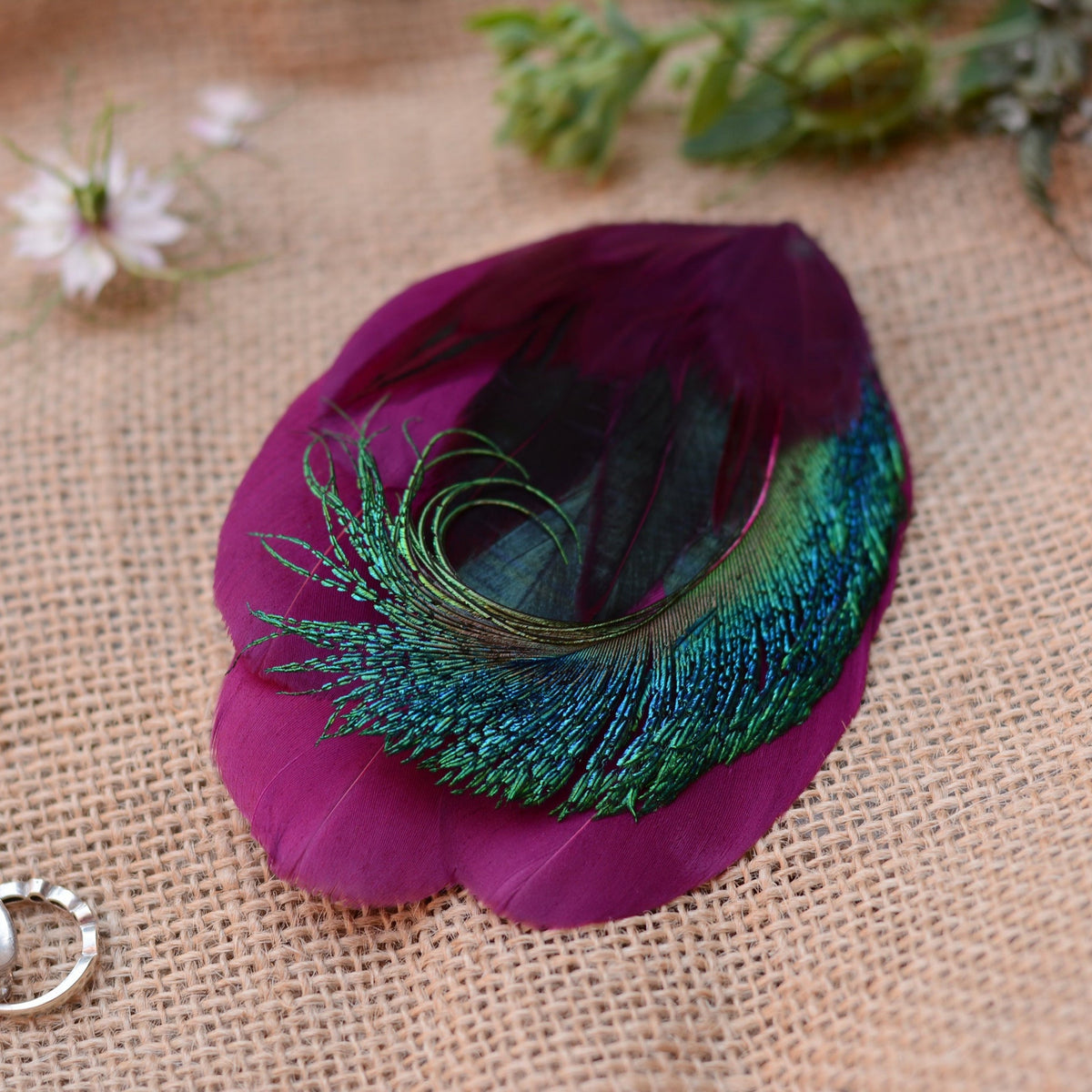 A decorative peacock feather with a vibrant purple body, shimmering teal-blue and green iridescence, and dark iridescent green inner feathers, displayed on a textured beige burlap surface with soft blurred flowers in the background.