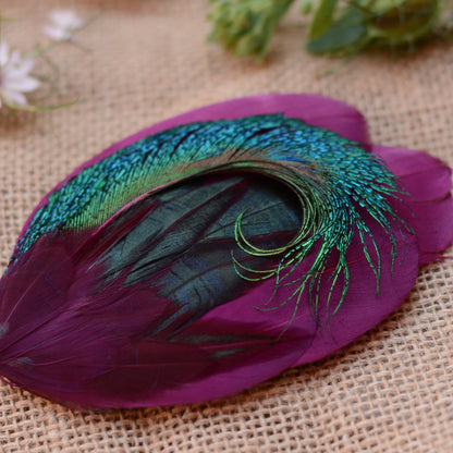 A decorative peacock feather with a vibrant purple body, shimmering teal-blue and green iridescence, and dark iridescent green inner feathers, displayed on a textured beige burlap surface with soft blurred flowers in the background.