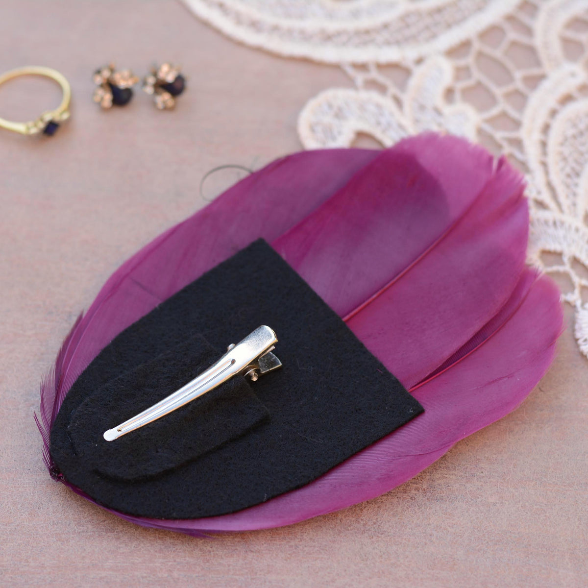 A feather hair clip place upside down to show a black felt base with a silver metal alligator style clip, displayed on a textured beige surface with lace and small jewelry props.