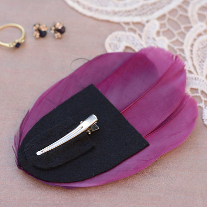 A feather hair clip place upside down to show a black felt base with a silver metal alligator style clip, displayed on a textured beige surface with lace and small jewelry props.