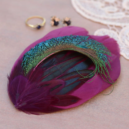 A decorative peacock feather with a vibrant purple body, shimmering teal-blue and green iridescence, and dark iridescent green inner feathers, styled on a beige surface with small jewellery props and lace in the background.