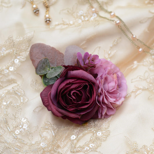 A close-up of a floral hair accessory featuring a deep burgundy rose, a light mauve flower, and green eucalyptus leaves, arranged on a cream lace fabric background with delicate jewellery.