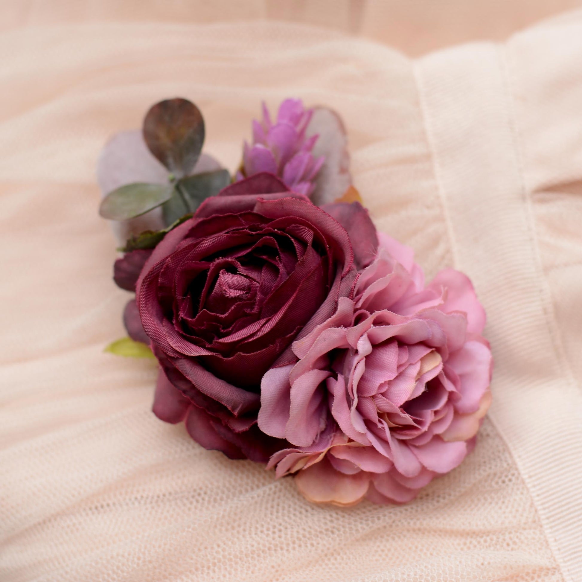 A close-up of a floral hair clip featuring a deep burgundy rose, a light pink peony, and green eucalyptus leaves, arranged on a soft beige fabric background.