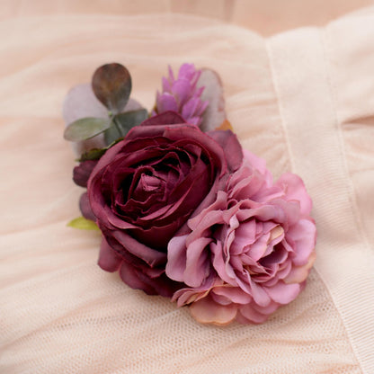 A close-up of a floral hair clip featuring a deep burgundy rose, a light pink peony, and green eucalyptus leaves, arranged on a soft beige fabric background.