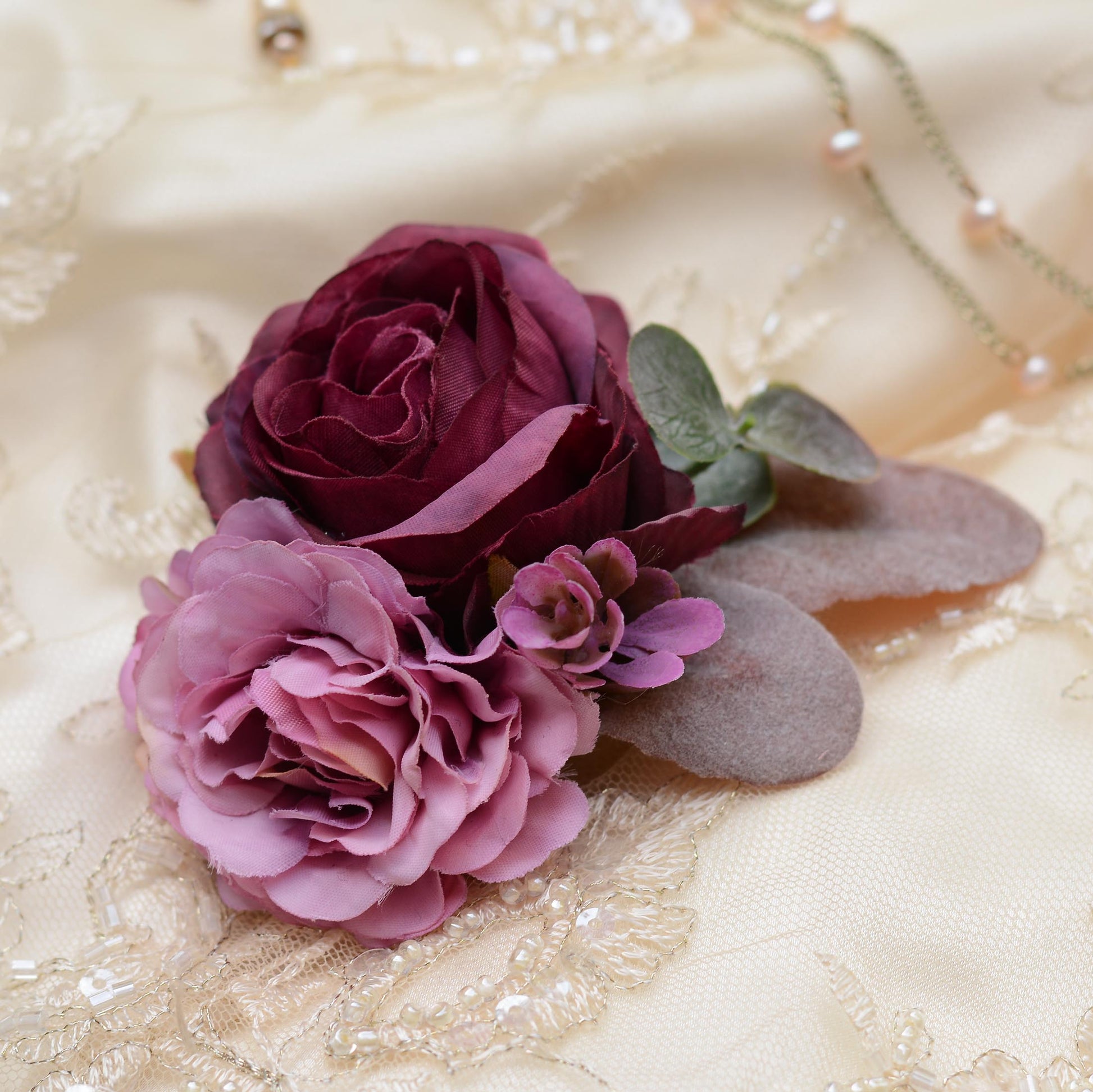 A close-up of a floral hair clip featuring a deep burgundy rose, a light mauve peony,  and dusty green eucalyptus leaves on a cream lace fabric background.