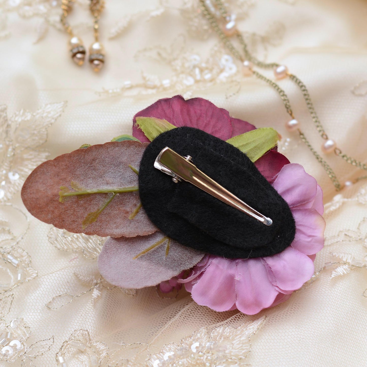 A floral hair clip with pink and purple fabric flowers, a dusty rose leaf, and a black felt center, with a silver alligator clip resting on a cream lace fabric background with gold necklaces.