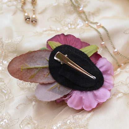A floral hair clip with pink and purple fabric flowers, a dusty rose leaf, and a black felt center, with a silver alligator clip resting on a cream lace fabric background with gold necklaces.
