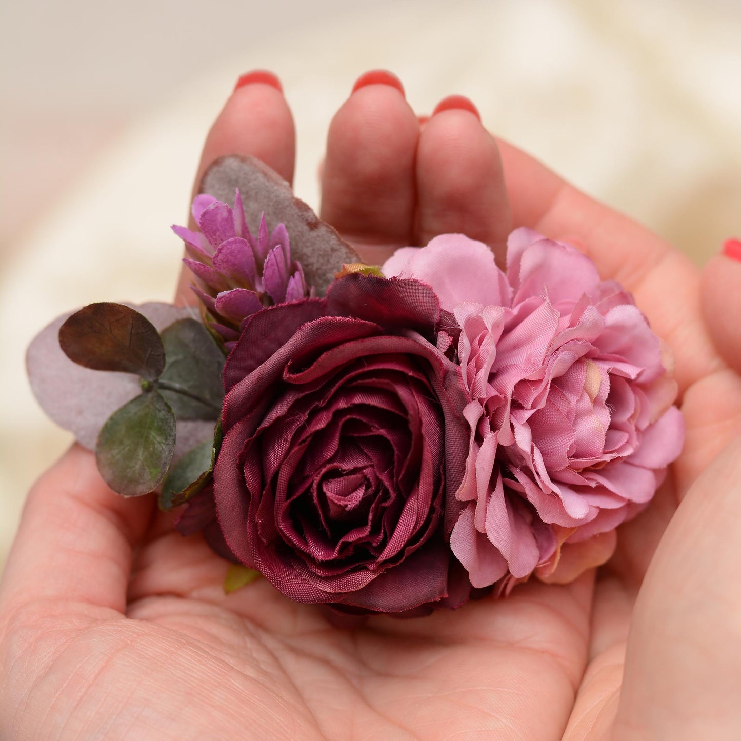 A close-up of a hand holding a floral hair clip with a deep burgundy rose, a mauve flower, and purple -green leaves against a soft cream background.