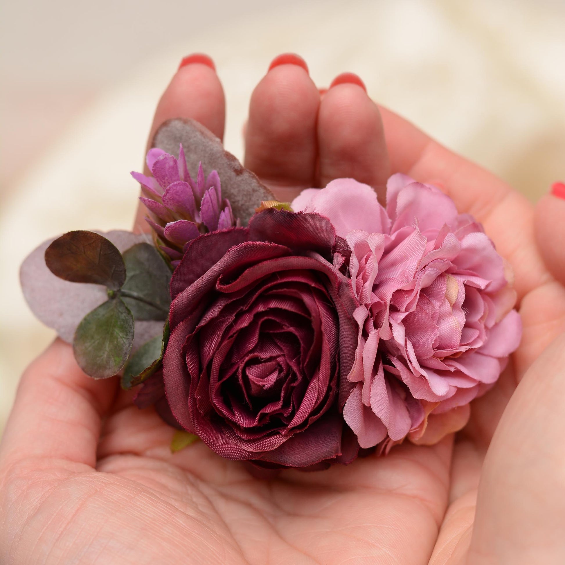 A close-up of a hand holding a floral hair clip with a deep burgundy rose, a mauve flower, and purple -green leaves against a soft cream background.