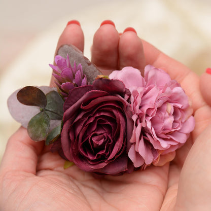 A close-up of a hand holding a floral hair clip with a deep burgundy rose, a mauve flower, and purple -green leaves against a soft cream background.