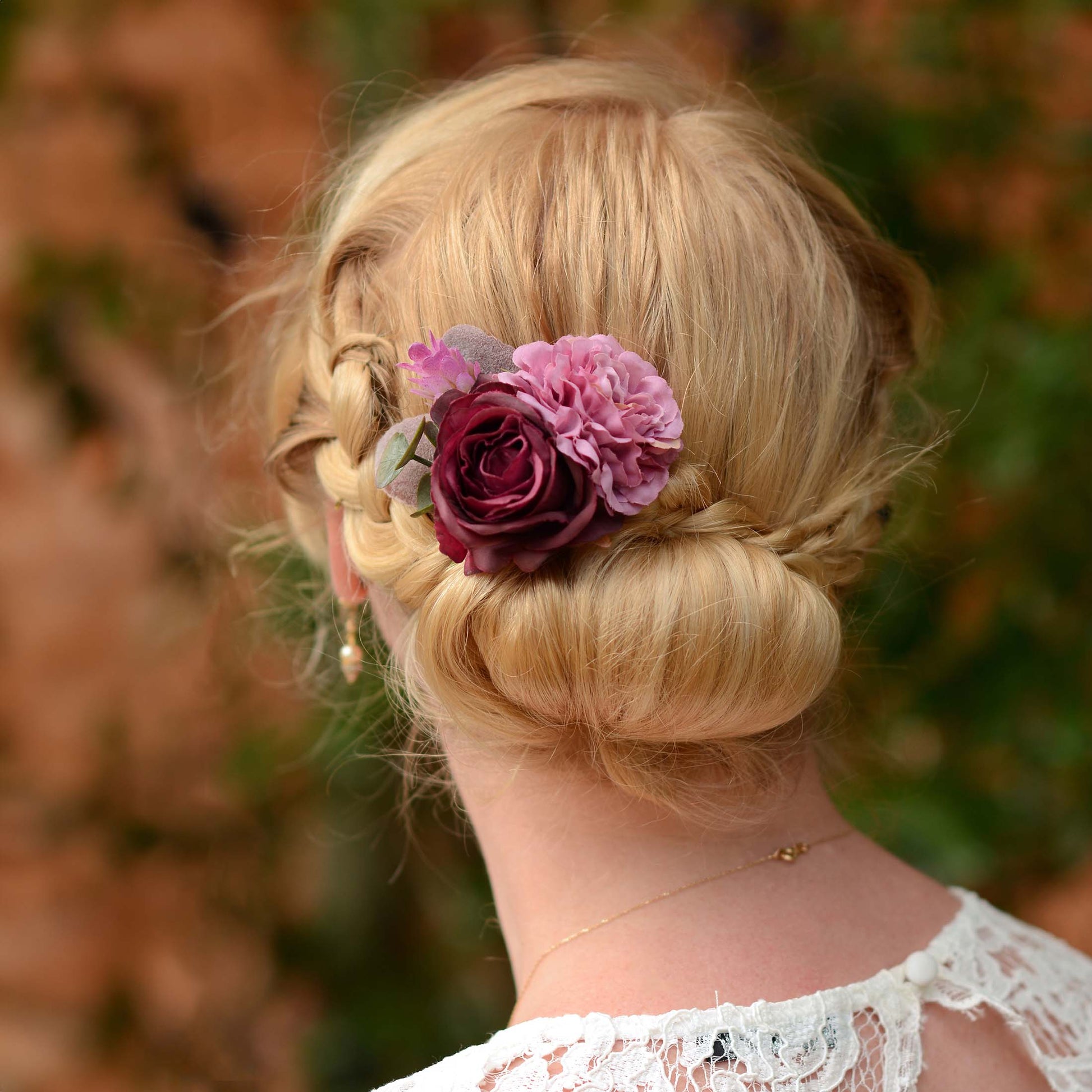 Blonde woman with braided hair and low bun decorated with a purple flower hair accessory against a blurred natural background