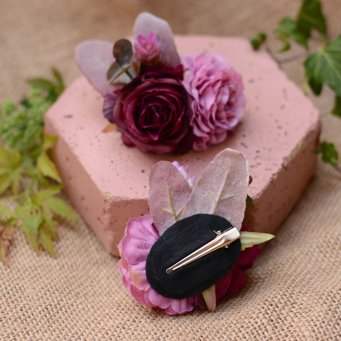 Hexagonal pink stone with purple and mauve flower hair accessories on a burlap cloth with green leaves.