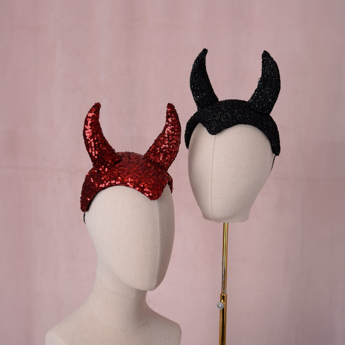 Two  devil horn headpieces displayed on mannequin heads against a soft pink backdrop. One headband is red sequined and the other is black glittery with two tall horns.