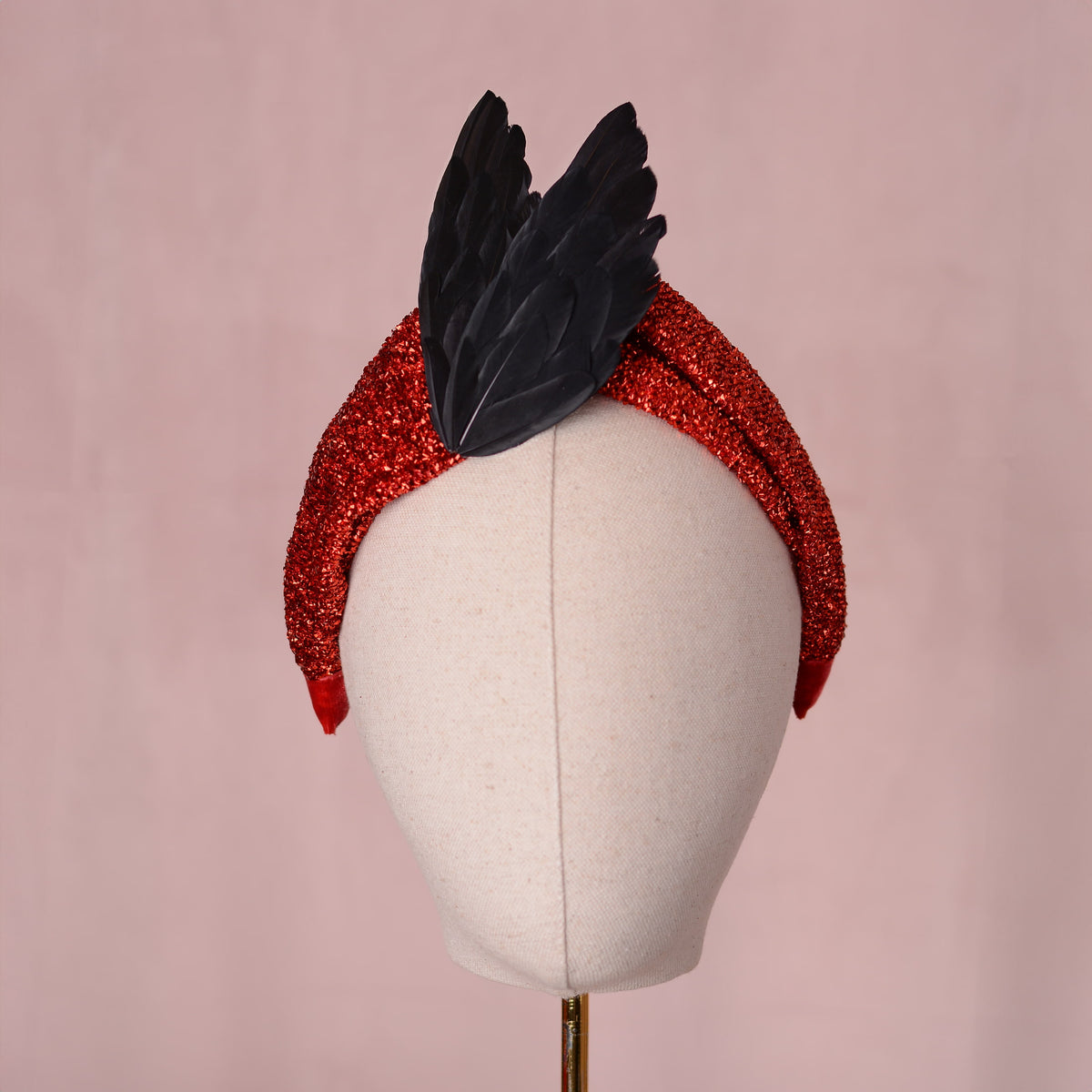 A red glitter headband with two black feather bird wings attached in an elgant v shape, displayed on a cream fabric mannequin head against a soft pink background.