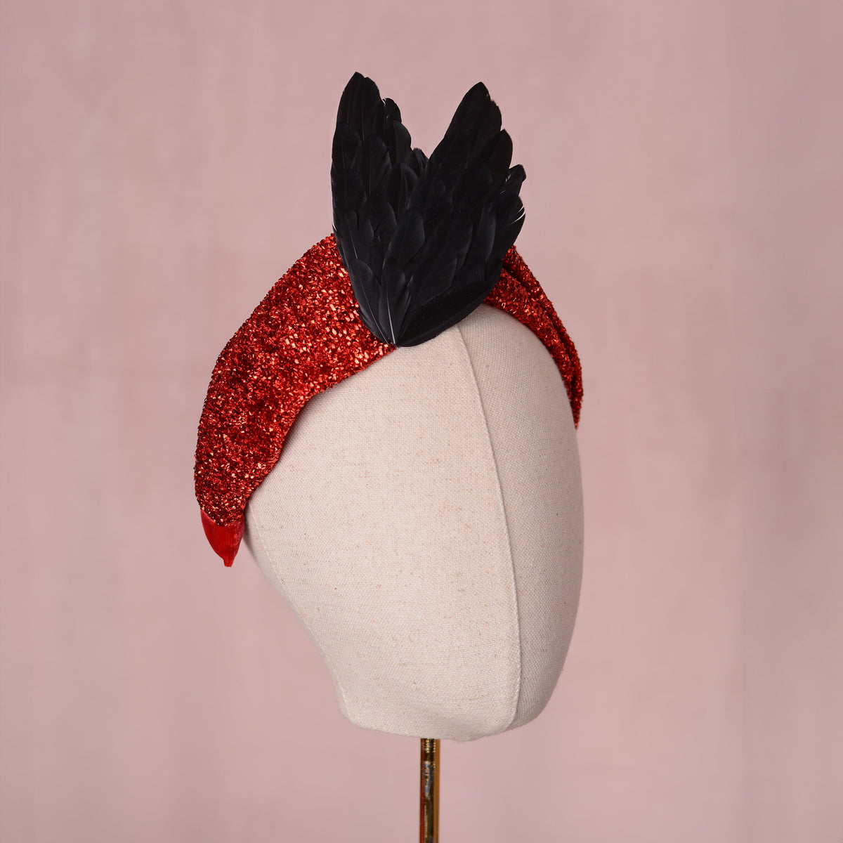 A red glitter fabric twist turban  headband with two large black feather bird wings attached at the top, displayed on a cream fabric mannequin head against a soft pink background.