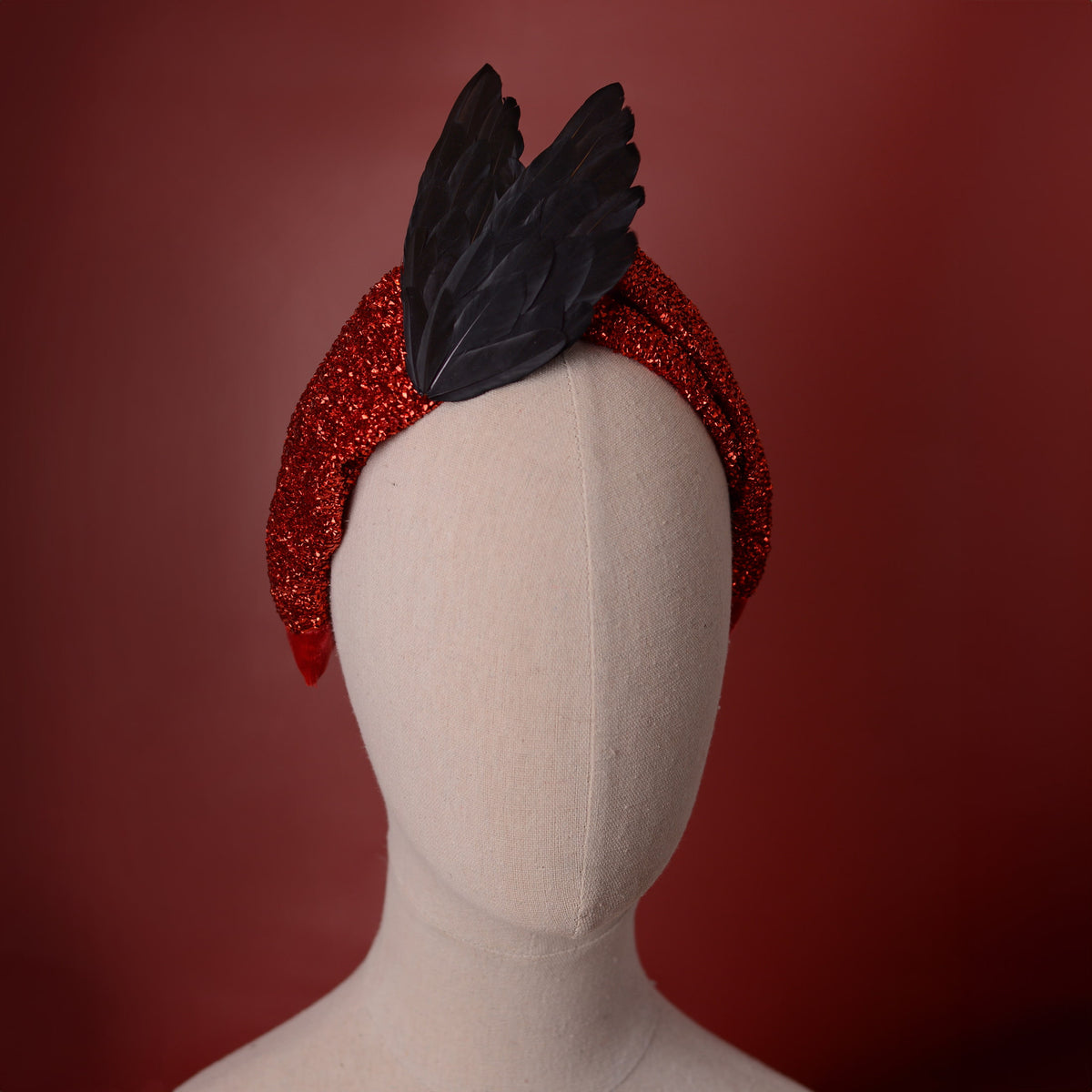Gothic vamp style red glitter twist turbanheadband with a black bird wing feather accent, displayed on a beige mannequin head against a dark red background.
