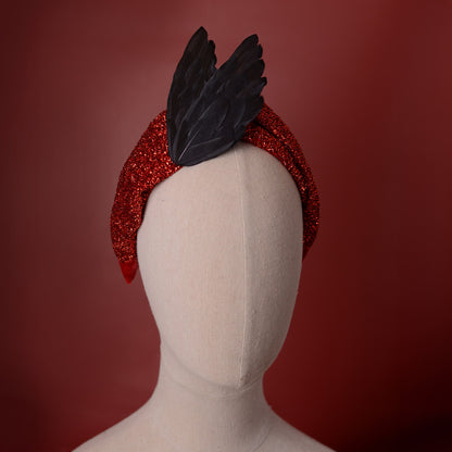 Gothic vamp style red glitter twist turbanheadband with a black bird wing feather accent, displayed on a beige mannequin head against a dark red background.