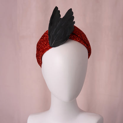 A red glitter headband styled with  black feather bird wings in an elegant v, displayed on a white mannequin head against a soft pink backdrop.