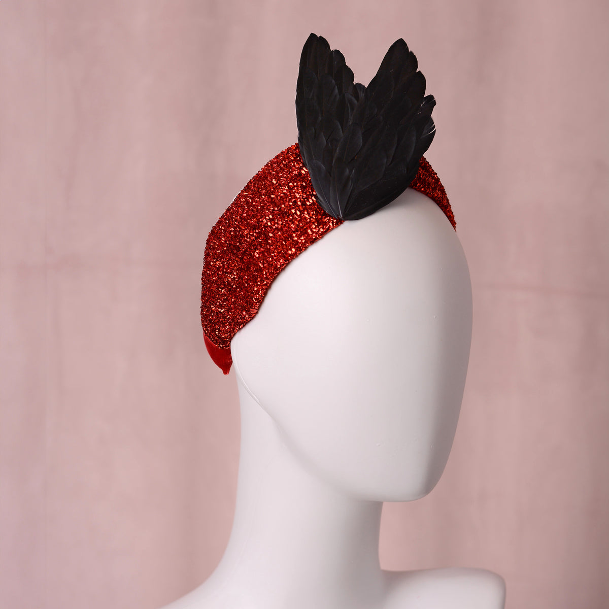 A red glitter headband with a black feather accent, displayed on a white mannequin head against a soft pink background.