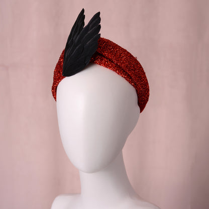 A red glittery headband styled on a white mannequin head, featuring a large black feather v shaped accent on top, set against a soft pink background. It has a gothic witchy vibe.