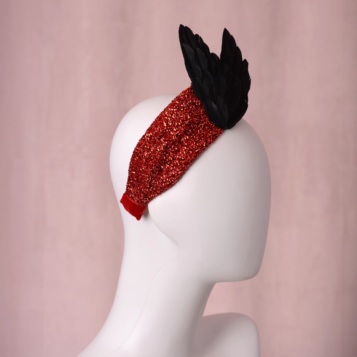 A red glitter fascinator headband with two black feather bird wing motifs displayed on a white mannequin head against a soft pink background.