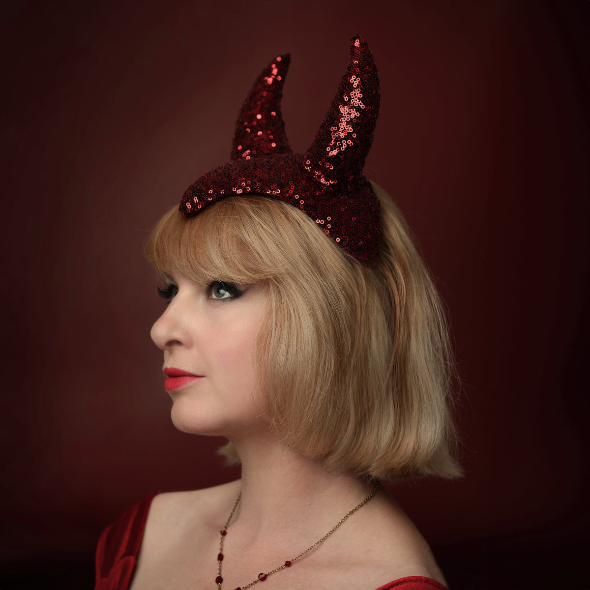 A woman with blonde hair wearing a red sequined devil horn headpiece in a retro showgirl style, shown in profile against a dark red background.