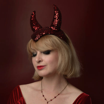 A woman with short blonde hair wearing a red sequined devil horn halloween party headpiece and a matching red velvet top, posed against a dark red background.