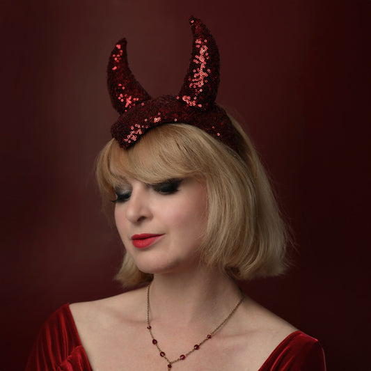 A woman with short blonde hair wearing a red sequined devil horn halloween party headpiece and a matching red velvet top, posed against a dark red background.