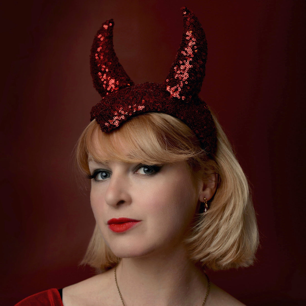 A woman with short blonde hair wearing a red sequined devil horn hat, posed against a dark red background.