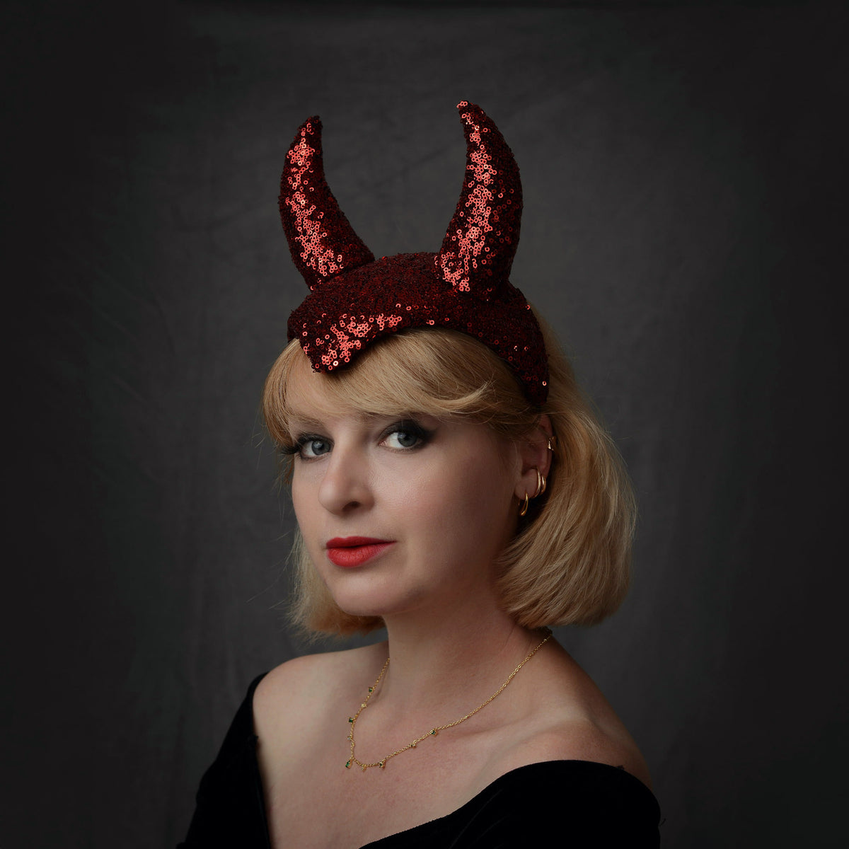 A woman with blonde hair wearing a theatrical red sequin devil horn headband, posing against a dark gray background.