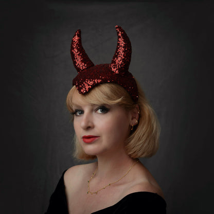 A woman with blonde hair wearing a theatrical red sequin devil horn headband, posing against a dark gray background.