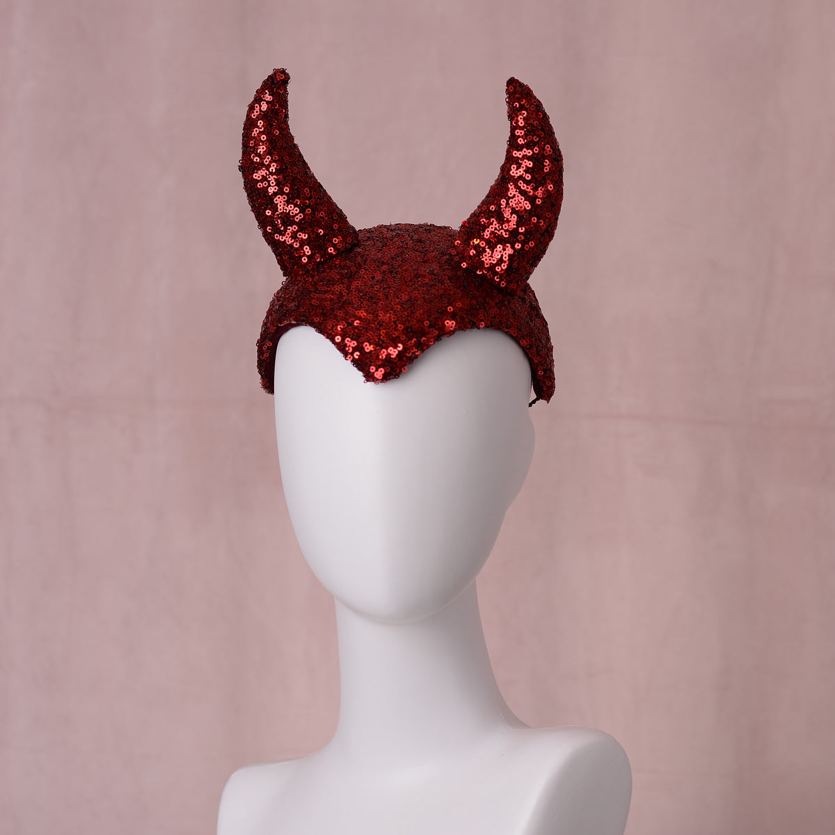 Red sequined theatrical red devil horn headband displayed on a white mannequin head against a soft pink backdrop.