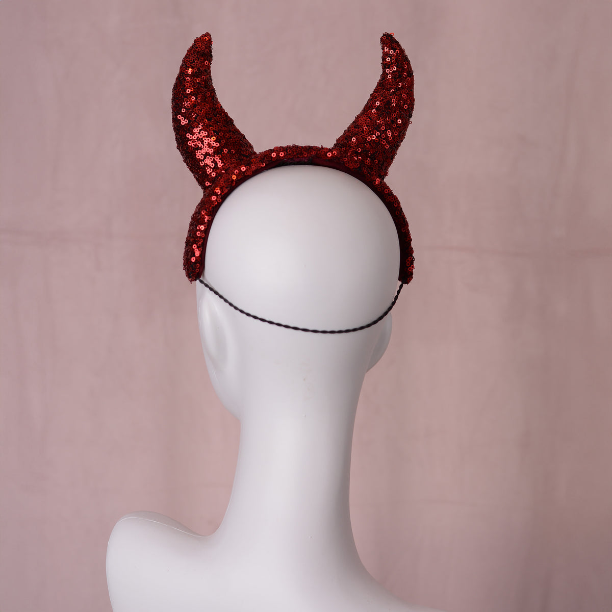 Red sequined devil horns headband with a black hat elastic, displayed on a white mannequin head against a soft pink backdrop.