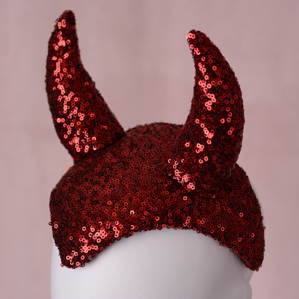 Red sequin devil horns costume headpiece displayed on a white mannequin head against a soft pink background.