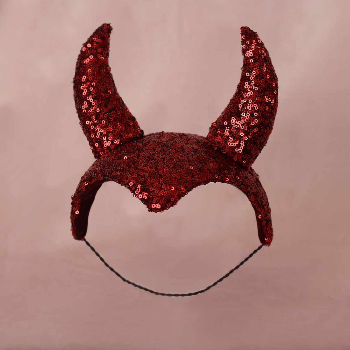 A red sequined devil horn headband headpiece with a black hat elastic, shown against a solid dusty pink background.