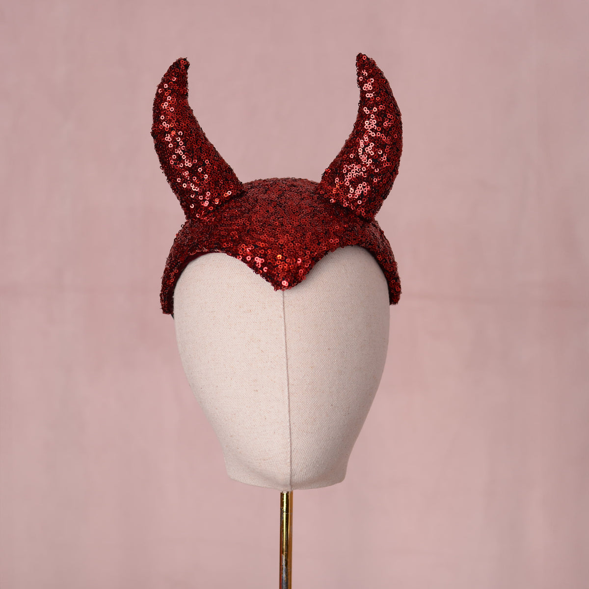 Red sequin devil horns headdress displayed on a white mannequin head against a soft pink background.