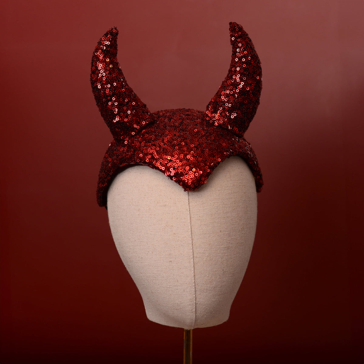 Red sequin devil horns show girl pin up style headpiece displayed on a beige fabric mannequin head against a dark red background.