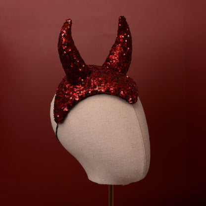 Red sequined devil horns headpiece displayed on a beige fabric mannequin head against a dark red background.