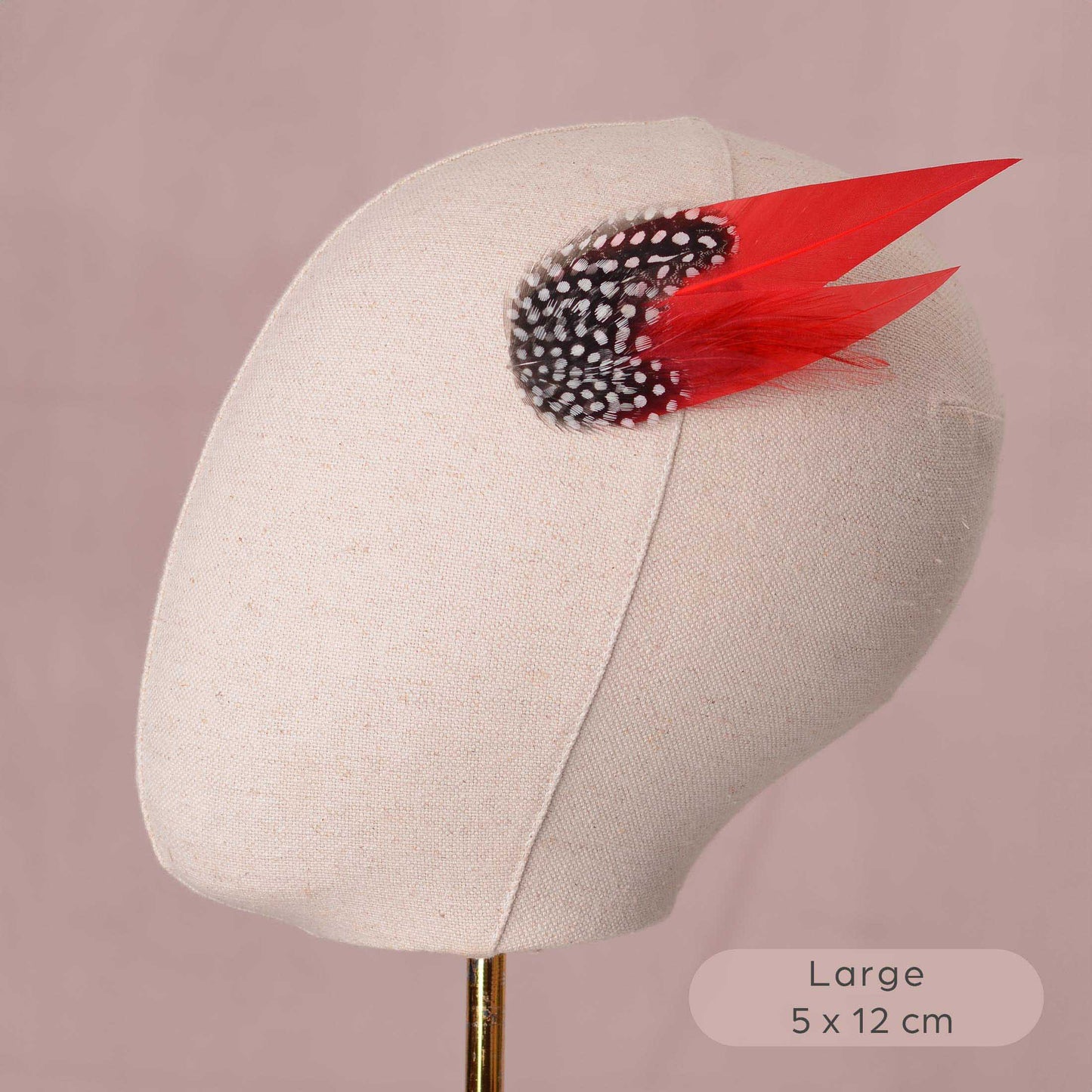 A beige fabric mannequin head display on a stand, featuring a decorative feather fascinator with a black and white speckled cluster and two bright red feathers, set against a dusty pink background with a size label in the corner.