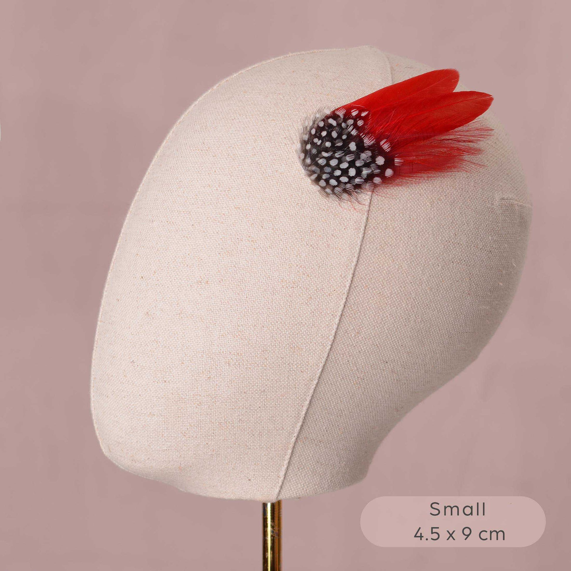 A small decorative hair accessory displayed on a beige fabric mannequin head against a dusty pink background. The accessory features two bright red feathers layered with  black and white polka dot feathers.