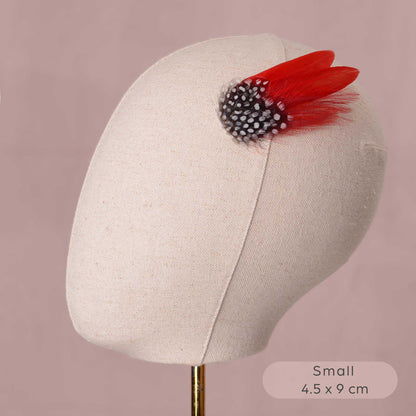 A small decorative hair accessory displayed on a beige fabric mannequin head against a dusty pink background. The accessory features two bright red feathers layered with  black and white polka dot feathers.