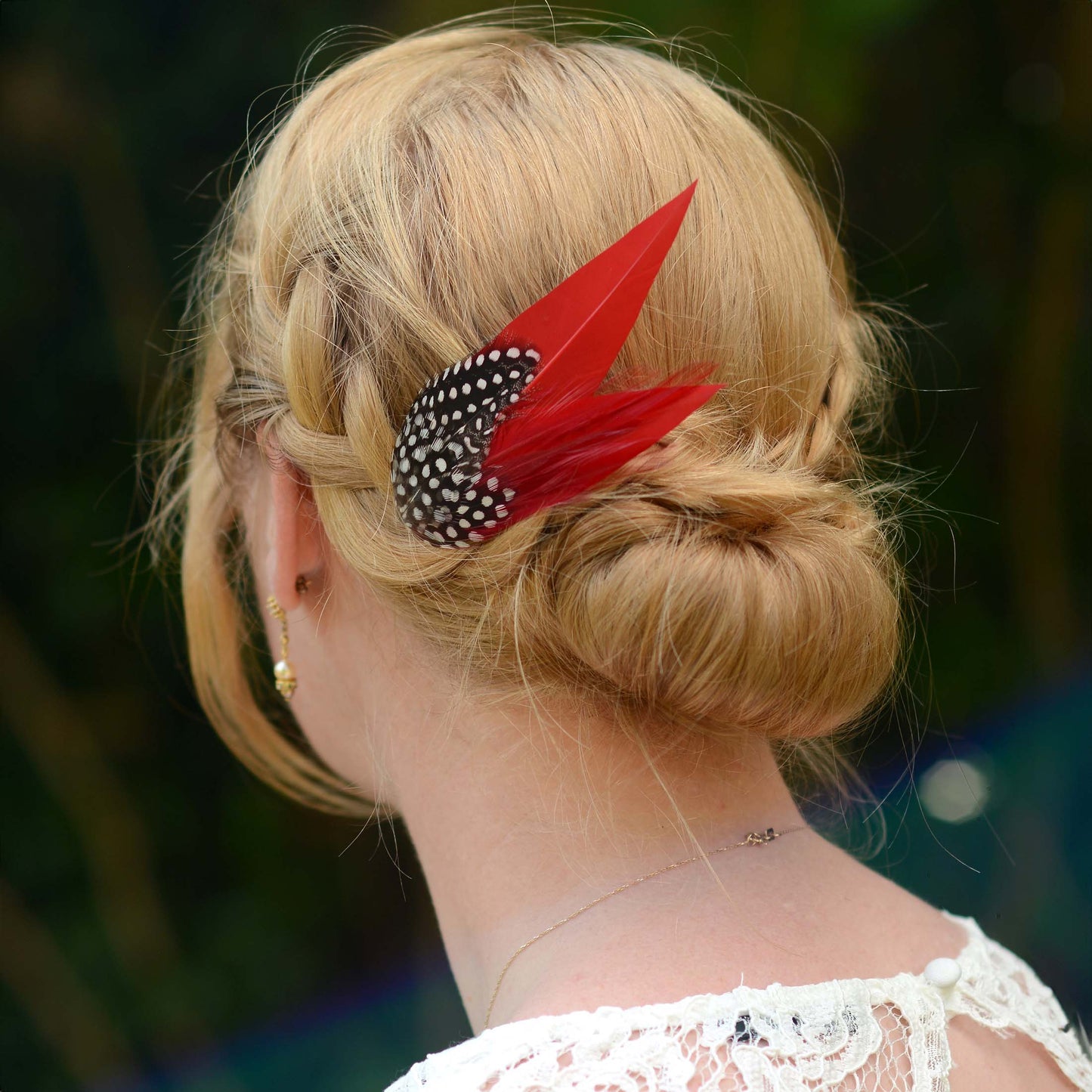 A blonde woman shown from the back wearing a red and black polka dot feather  hair clip accessory in her low bun, with a blurred outdoor background.