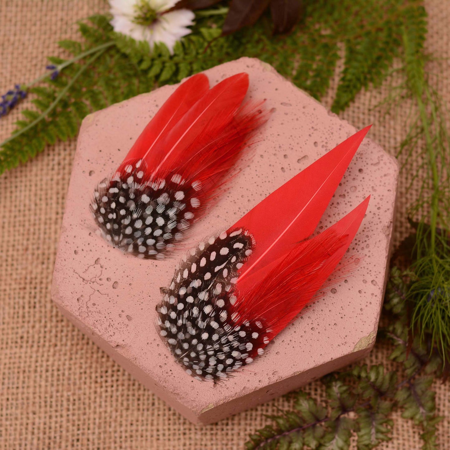 Two red-and-black-and-white spotted feather hair accessories displayed on a pink hexagonal stone, surrounded by burlap fabric, green leaves, and a white flower.