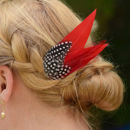A close-up of a blonde woman's braided hair styled in a low bun, featuring a decorative hair accessory with two bright red feathers and black-and-white spotted feathers.