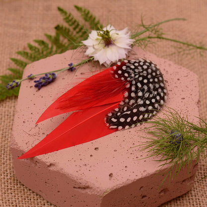 A decorative feather  hair ornament with black and white polka dot feathers, on pointed red feathers, displayed on a pink stone with flowers and greenery.