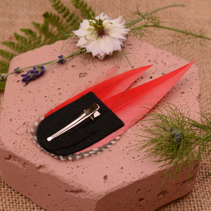 A decorative red  hair clip styled on a pink stone slab with botanical props. The clip features two bright red feathers, a black felt base, and a silver metal alligator clip mechanism.