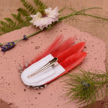 A decorative red  hair clip styled on a pink stone slab with botanical props. The clip features two bright red feathers, a white felt base, and a silver metal alligator clip mechanism.