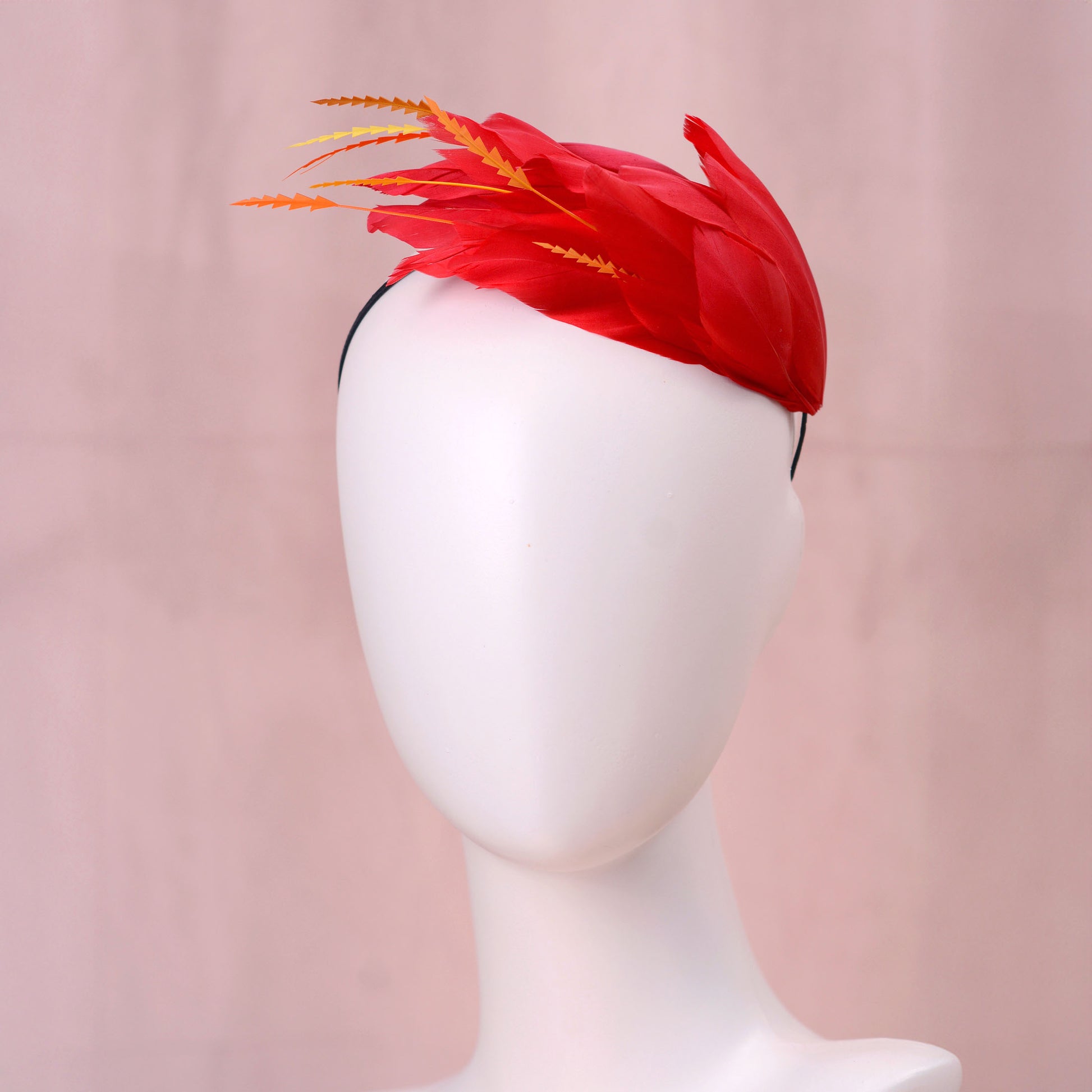 A bright red feather fascinator hat with orange-tipped feathers, displayed on a white mannequin head against a soft pink background.